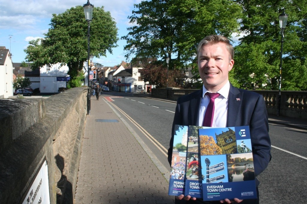 Cllr Bradley Thomas holding three documents stood on a foot bridge with the town in the background