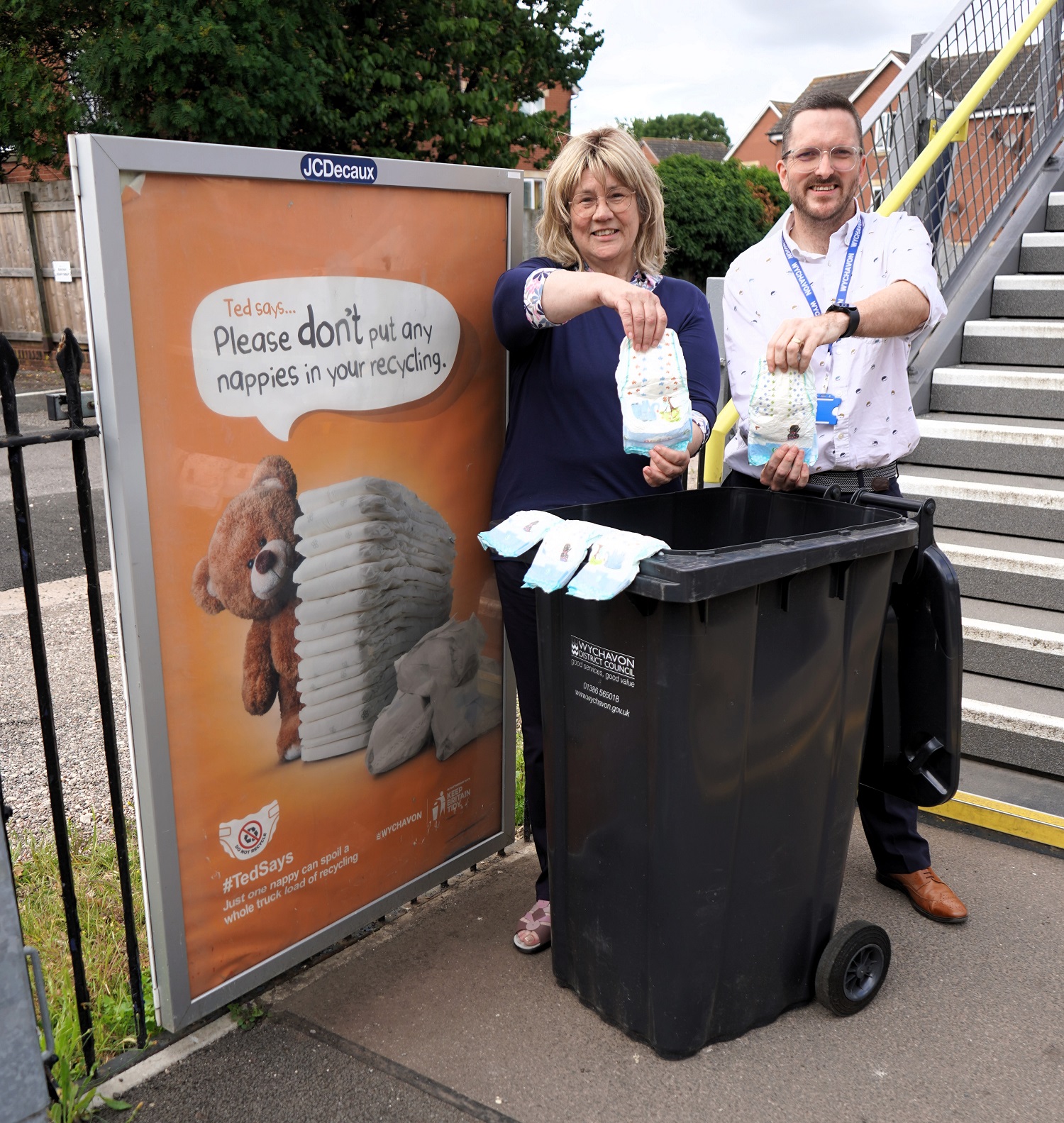 A man and a woman holding disposable nappies over a black bin in front of a poster