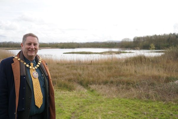 A man stood in front of a lake and some grass fields.