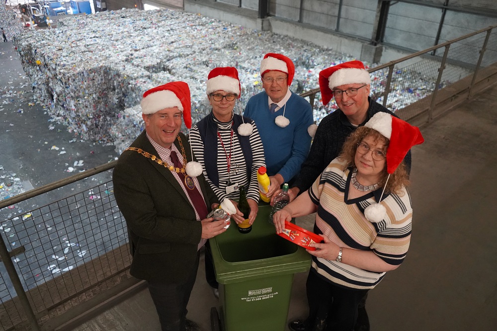 A group of people stood around a green recycling bin with Christmas hats on holding recycling.
