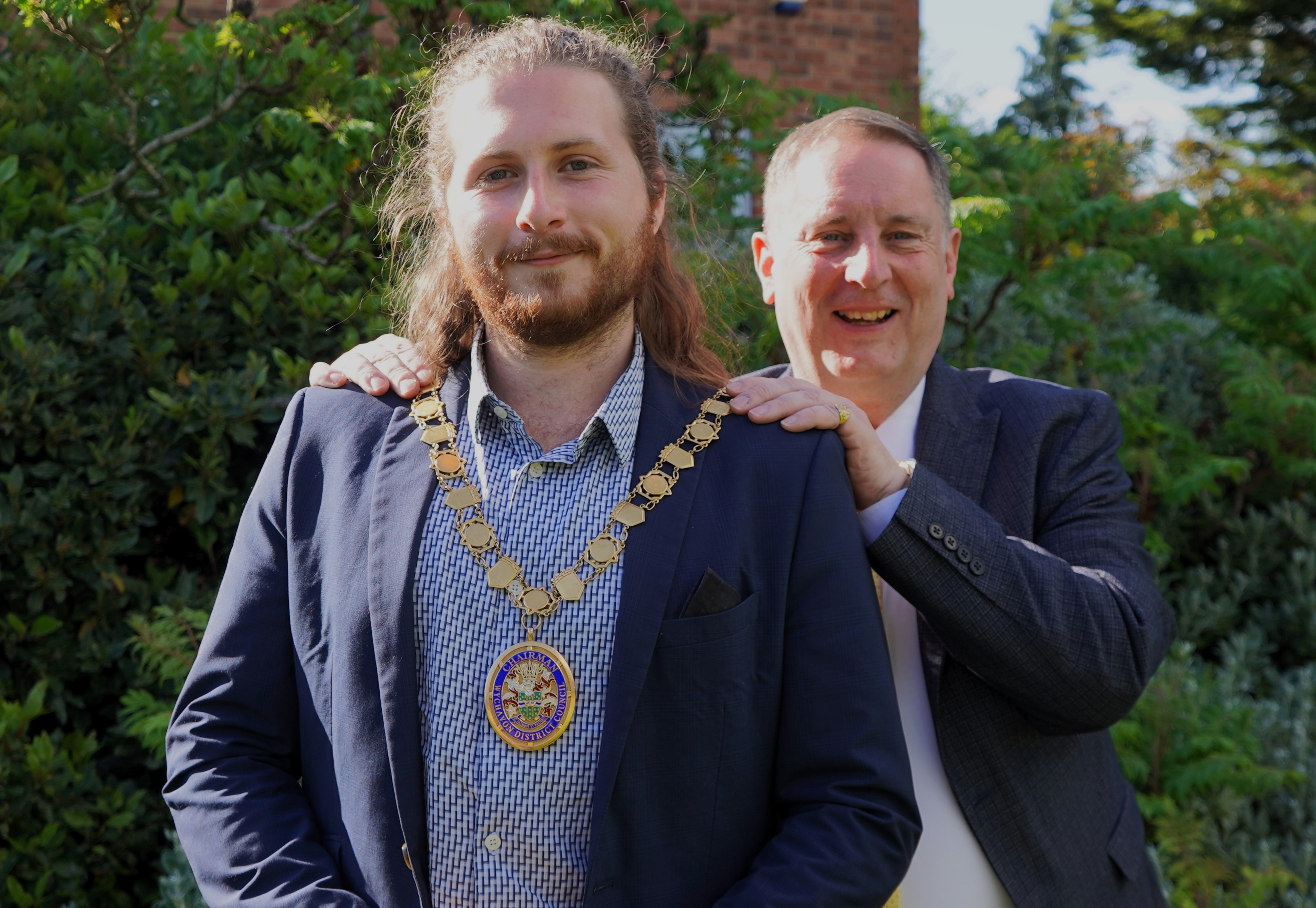 A man stood behind another man putting a mayoral chain over his head.