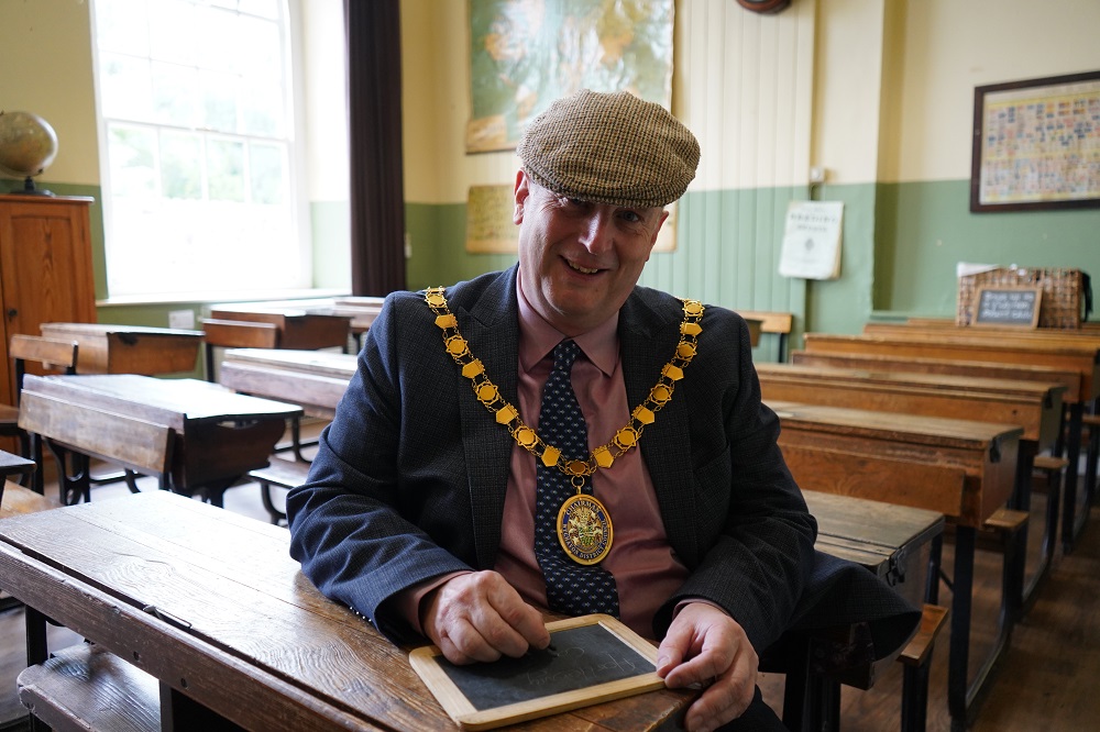 Cllr Raphael wearing a flat cap hat writing on a chalk slate board behind and old school desk.