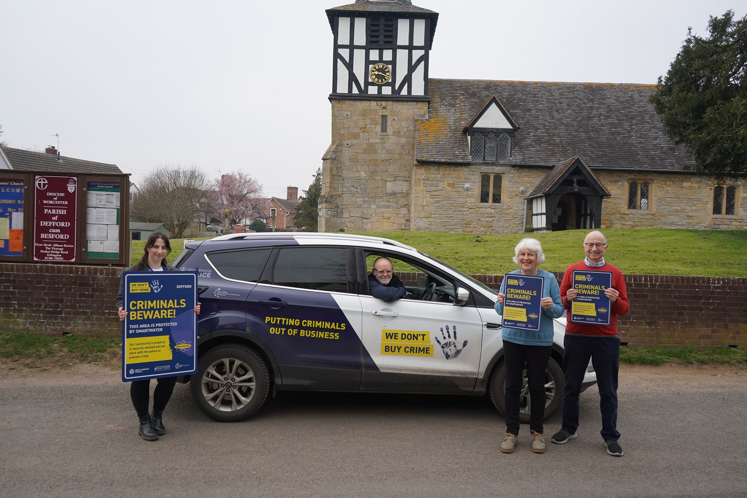 A car in front of a church. A man leans out the window. Three people are stood either side holding Criminals beware signs.