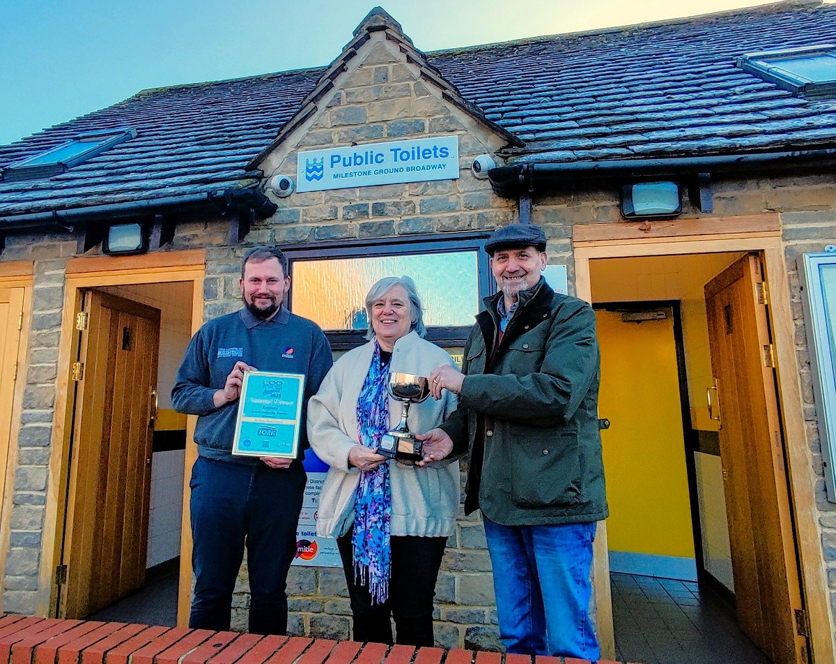 Three people stood in front of public loos holding a certificate and a trophy.