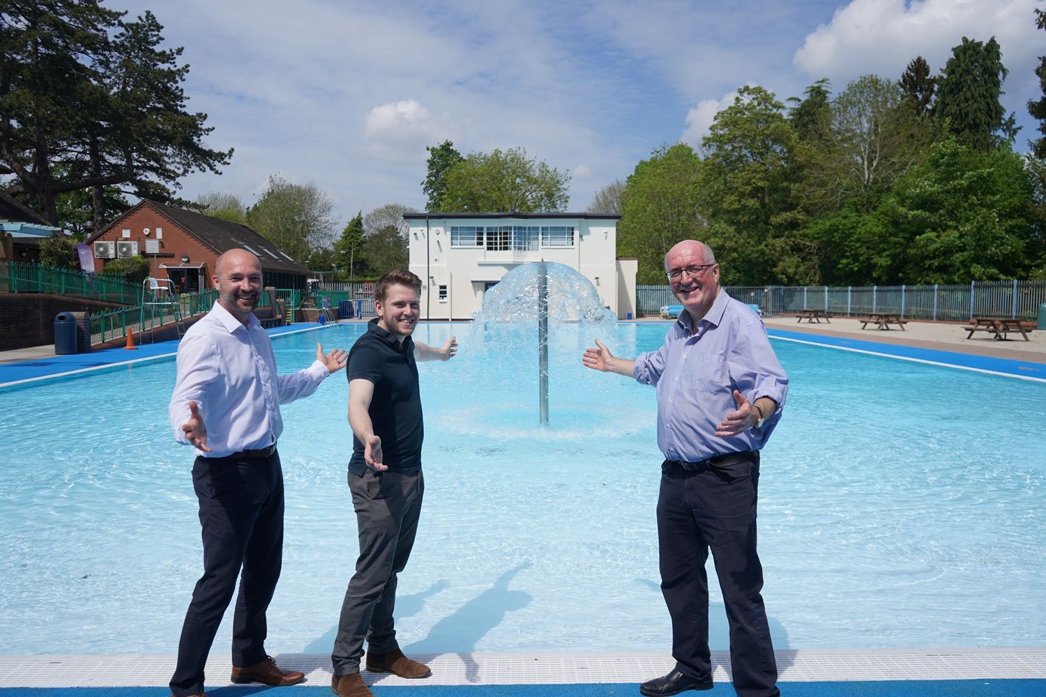 Three men stood in front of an outdoor swimming pool holding information boards.