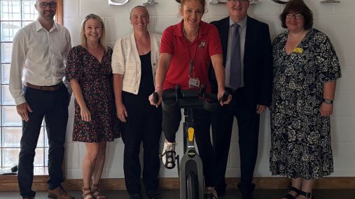 A woman on an exercise bike at the front of the picture with five people stood either side in the background in a line