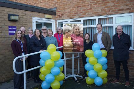 A group of people stood in front of some balloons and either side of a banner with some smiling people and Defy Dementia on it.
